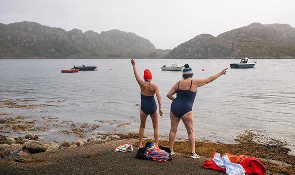 Two mature women in bathing suits standing on lakeshore facing the lake.